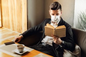 Handsome adult bearded man indoors in cafe. Lifestyle concept photo with copy space. Picture with book and protective face mask
